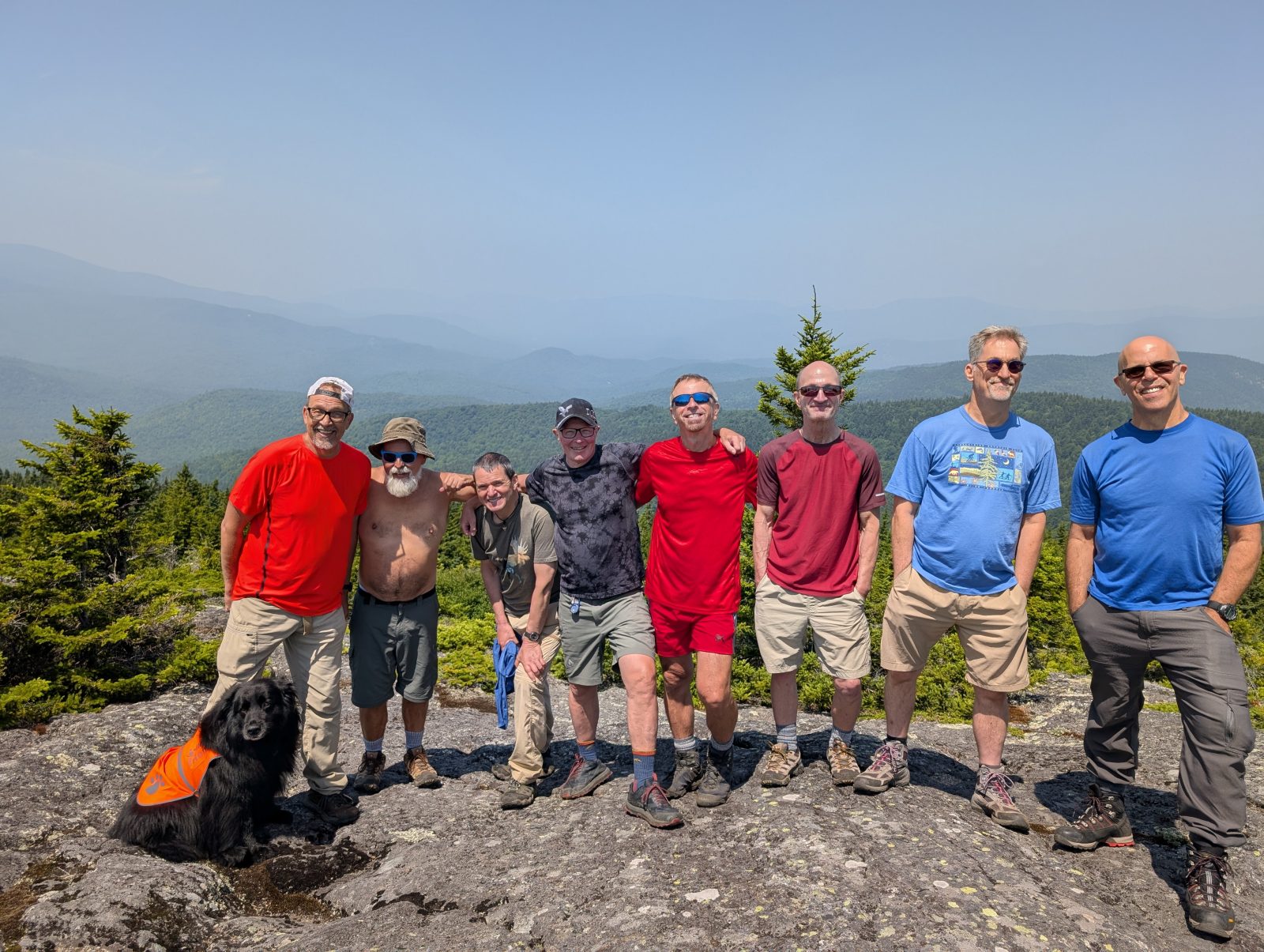 GayOutdoors members and a dog on a summit ledge with mountain ridges behind