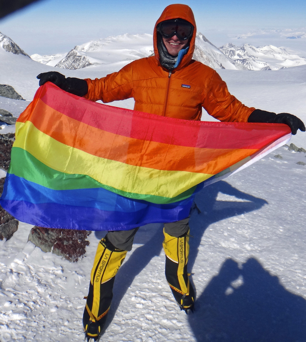 A climber holding a rainbow pride flag on a snowy mountain summit